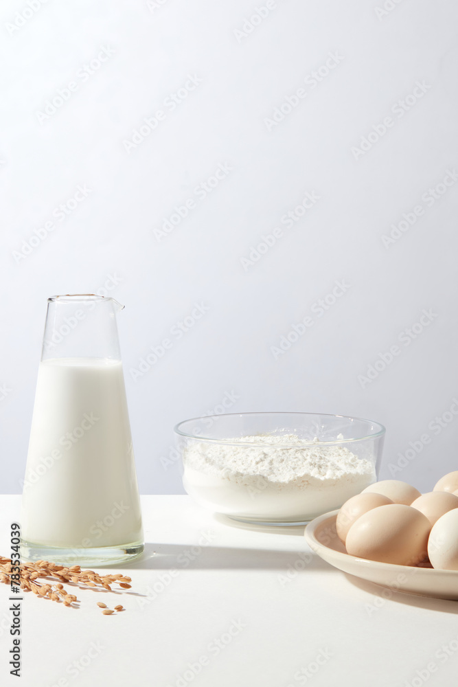 Milk filled in conical glass bottle without top, a glass bowl of flour and some eggs contained in white round dish on minimals white background. Photo for product of cooking