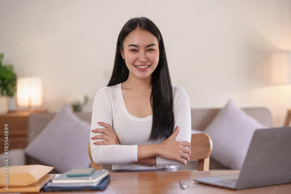 Confident young asian woman smiling with arms crossed in home office.