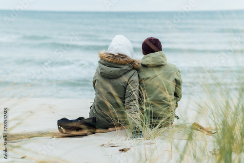 Fototapeta Naklejka Na Ścianę i Meble -  A couple in love sits on the beach, dreams and looks at the sea. Rear view, against the backdrop of the sea and waves