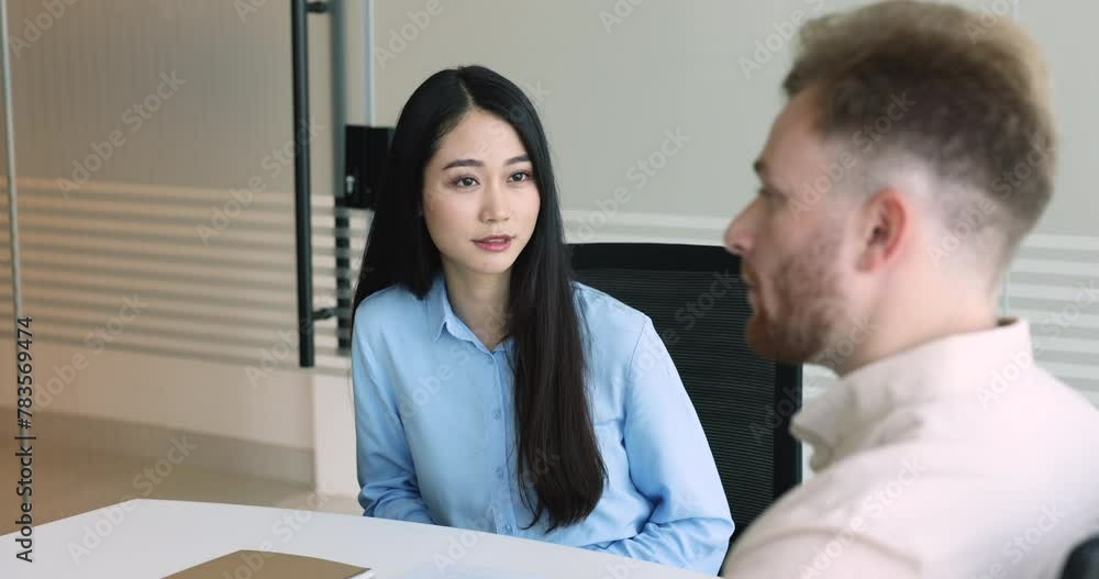 Positive young Asian new employee woman talking to mentor, listening to boss, male colleague, reviewing notebook, smiling. Two diverse coworkers discussing project at workplace table Positive young Asian new employee woman talking to mentor, listening to boss, male colleague, reviewing notebook, smiling. Two diverse coworkers discussing project at workplace table