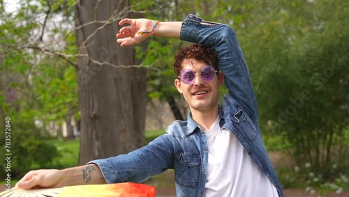 Happy caucasian casual gay man dancing with rainbow fan in a park photo