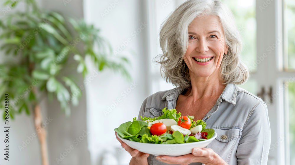 A happy elderly woman holding a bowl of fresh organic green vegetable salad in a light kitchen. Eating healthy food. Concept of Diet, Fitness, Healthy lifestyle, vegetarianism. A happy elderly woman holding a bowl of fresh organic green vegetable salad in a light kitchen. Eating healthy food. Concept of Diet, Fitness, Healthy lifestyle, vegetarianism.