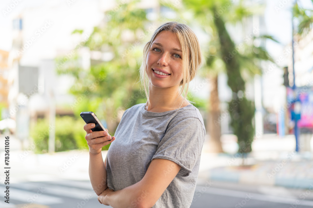 Young blonde woman at outdoors holding a mobile phone and with arms crossed Young blonde woman at outdoors holding a mobile phone and with arms crossed