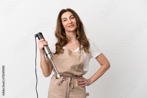 Middle aged caucasian woman using hand blender isolated on white background posing with arms at hip and smiling