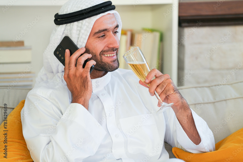 Relaxed Arabian man in a Keffiyeh talking on the phone with a content smile, casually holding a flute of bubbly in a modern setting. Relaxed Arabian man in a Keffiyeh talking on the phone with a content smile, casually holding a flute of bubbly in a modern setting.