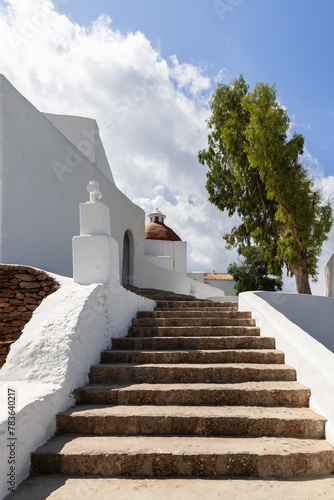 The tranquil setting of Puig de Missa church in Ibiza  Spain  captured in a vertical frame. White walls contrast with the brown steps and terracotta dome  complemented by a lush tree and blue sky