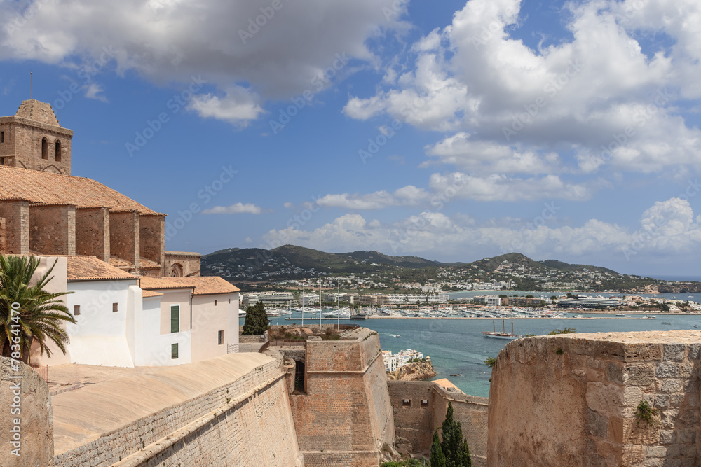 The historic walls of Eivissa Castle frame a stunning view of Ibiza bustling port, blending the old with the new amid serene blue waters and rolling hills under a dynamic sky