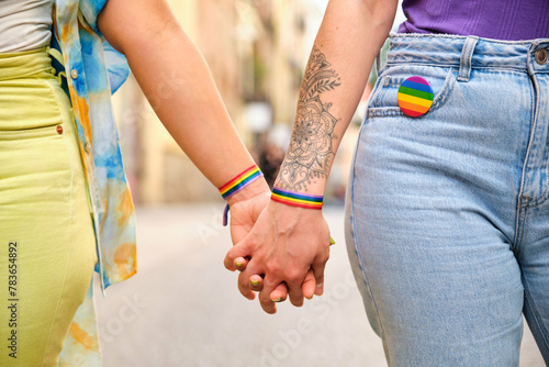Unrecognizable lesbian couple holding hands with LGBTQ flag bracelets and pin. Pride Parade celebration.