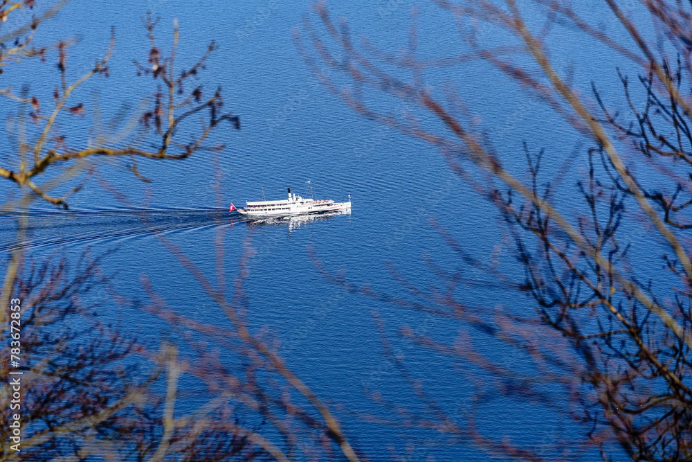 Naklejka premium Aerial view of white paddle steamer Uri with waving Swiss flag crossing Swiss Lake Lucerne on a sunny spring day. Photo taken April 11th, 2024, Bürgenstock, Lake Lucerne, Switzerland.