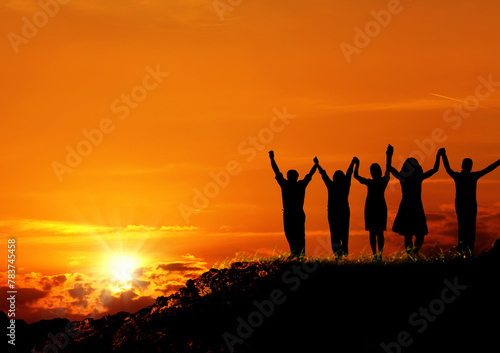 group of silhoutted people on hill arms raised and holding hands over orange colored sunset sky