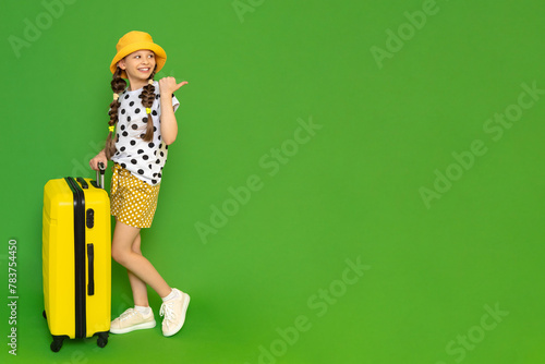 A little girl with a suitcase on wheels is looking at your advertisement. A child in shorts and a hat is resting for the summer. Green isolated background. Copy space.