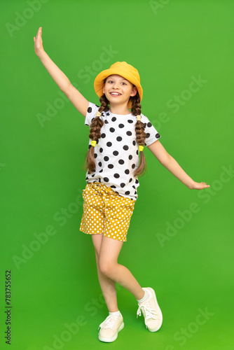 A little girl in a hat and shorts is enjoying the warm summer. Children's summer holidays.  The young girl opens her arms in different directions and smiles broadly. Green isolated background.