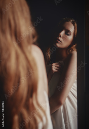 Dramatic portrait of beautiful young sad girl among the dark. Sorrowful woman stands at the mirror