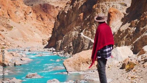 Rear view shot of a defocused Indian man wearing hat and shawl looking at the blue Tsarap Chu river on the way towards Phugtal Monastery in remote area of Zanskar Valley in Ladakh, India.  photo