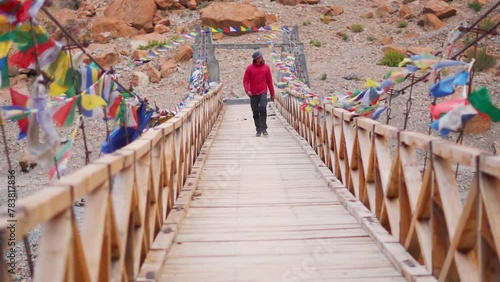 Indian male tourist wearing hat walking on the wooden bridge above the Tsarap Chu river on the way towards Phugtal Monastery at Zanskar Valley in remote regions of Ladakh, India. Tourist on bridge. photo