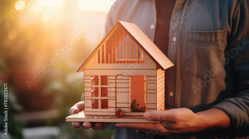 Man holding wooden house model against sunset light for sale or rent, family home and shelter concept, 