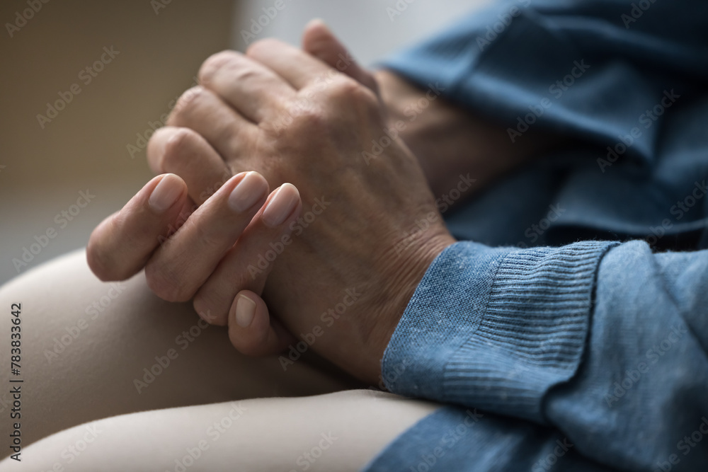 Fototapeta premium Cropped shot unknown older woman sit on sofa, folded palms close up view. Experiencing stressful situation, news about disease, suffering from loneliness or illness. Disorder, nursing house, solitude