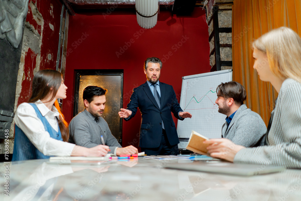 Office workers sit in a conference room with red walls and the director explains the drawn up schedule