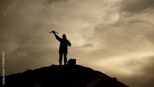 A solitary soldier's silhouette stands out on a hilltop as he launches a drone into the twilight sky, with the dramatic backdrop of a vibrant sunset providing a powerful contrast. Camera 8K RAW.