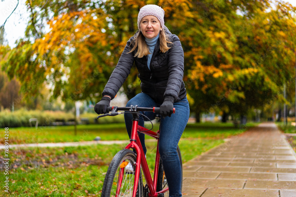 Obraz premium Mid-adult woman riding bicycle in city park on a rainy day 