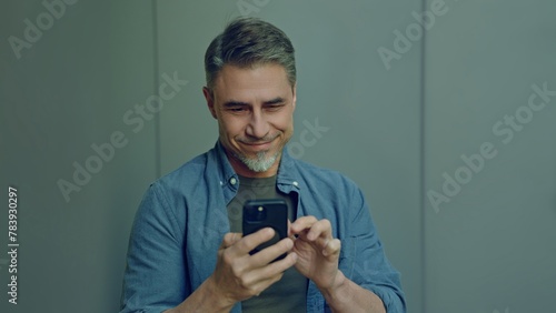 Portrait of a content middle-aged man looking at cell phone. Happy, confident mid adult male in casual. Blank copy space on a gray wall background.