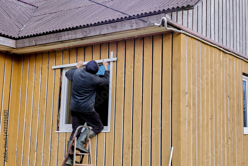 Standing on steel folding stepladder, construction worker installs external slopes on window of rural house from facade.