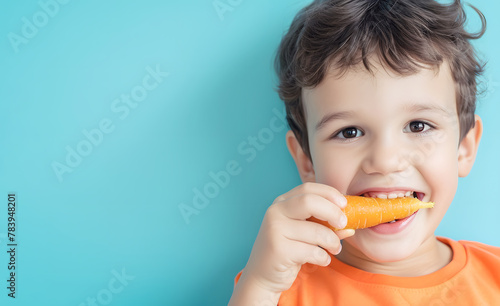 Toddler boy eats fresh carrot on blue background closeup. Happy child bites juicy vegetable in studio. Healthy ration for little kids concept