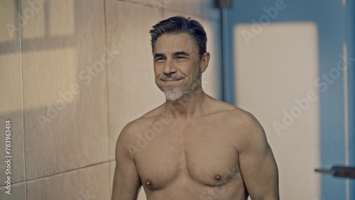 Confident mature male standing shirtless in front of a bathroom mirror. Portrait of muscular fit aged man, smiling.