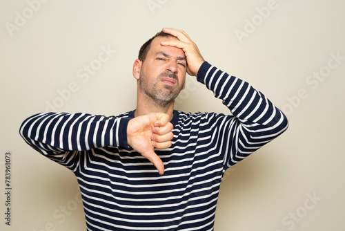 Hispanic man with beard about 40 years old wearing striped sweater looking unhappy and angry showing rejection and negative with thumbs down gesture. isolated on beige studio background.