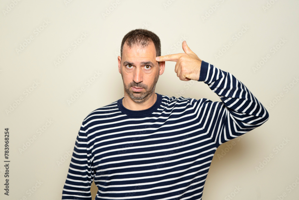 Hispanic man with beard in his 40s wearing a striped sweater standing over a beige background shooting and killing himself pointing with hand and fingers at his head like a gun, suicidal gesture Hispanic man with beard in his 40s wearing a striped sweater standing over a beige background shooting and killing himself pointing with hand and fingers at his head like a gun, suicidal gesture