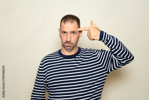 Hispanic man with beard in his 40s wearing a striped sweater standing over a beige background shooting and killing himself pointing with hand and fingers at his head like a gun, suicidal gesture