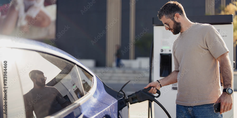 Adult male plugs in an electric vehicle for charging at a modern charging point during a sunny day. Adult male plugs in an electric vehicle for charging at a modern charging point during a sunny day.