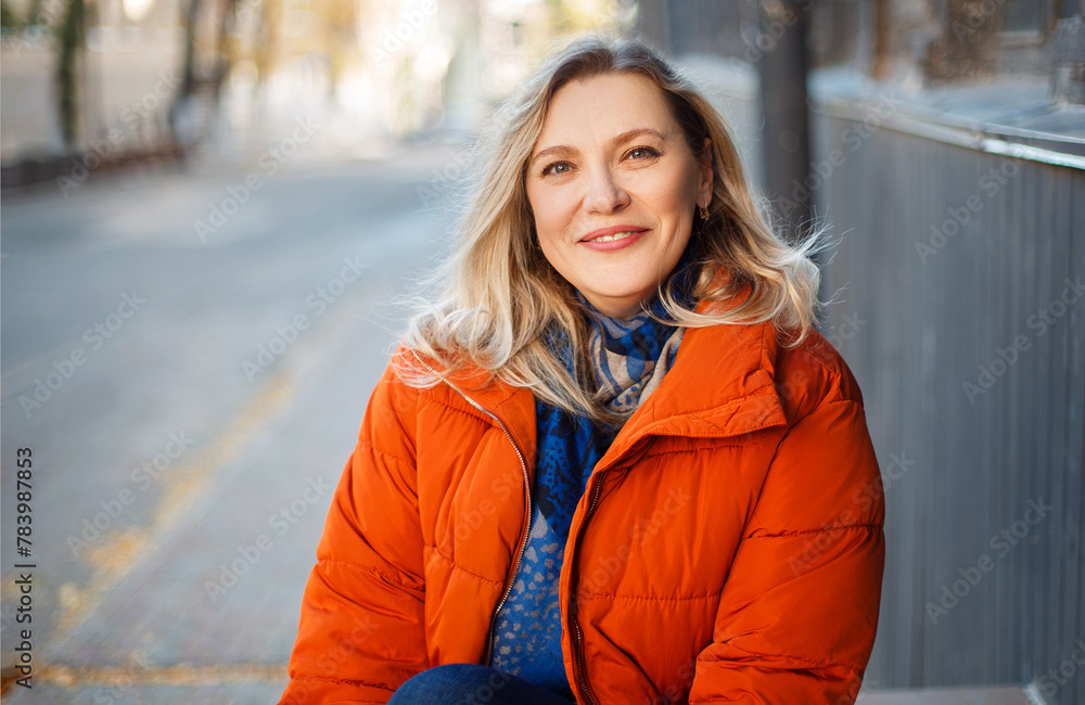 Happy smiling middle aged woman in orange down jacket sitting on concrete stairs outdoors