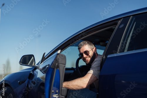 Portrait of a cheerful, bearded man wearing sunglasses as he leans out of his new electric car, enjoying a sunny day with clear blue skies.