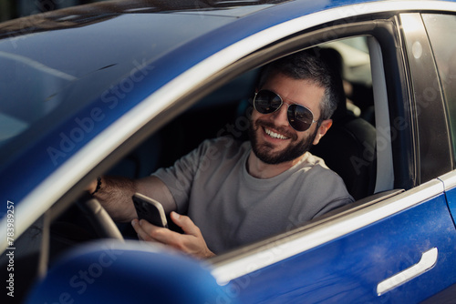 Smiling man with sunglasses using his smartphone while sitting in the driver's seat of a modern electric car. © arthurhidden