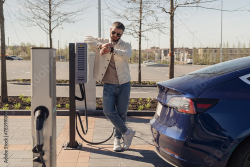A stylish adult male checks his watch as he waits by his electric car, which is charging at a modern charging station on a sunny day.