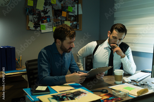 Two focused detectives review documents at a cluttered investigation bureau desk