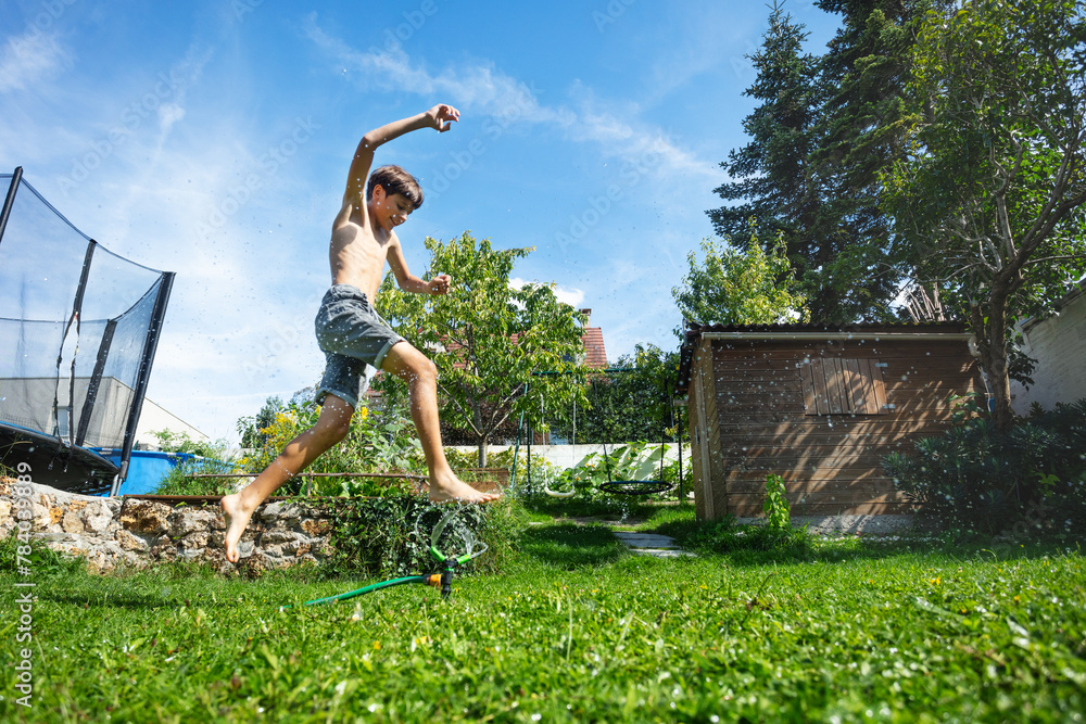 Boy bursts with energy as he vaults a spray of water in backyard - obrazy, fototapety, plakaty 