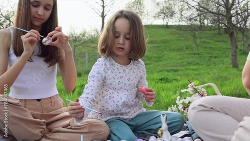 a family of three girls of different ages and European appearance are sitting in the garden on the green grass and are engaged in painting Easter eggs. Easter traditions. Spring holiday.