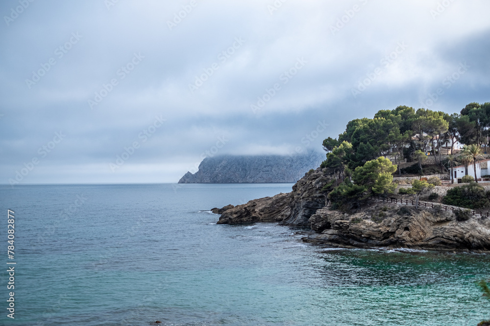 Misty seascape with a rocky coastline, the sea meeting a cloud covered mountain under a subdued sky.