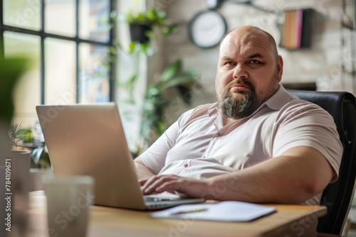 Adult businessman with beard looks seriously at the camera while working on his laptop in a well-lit office