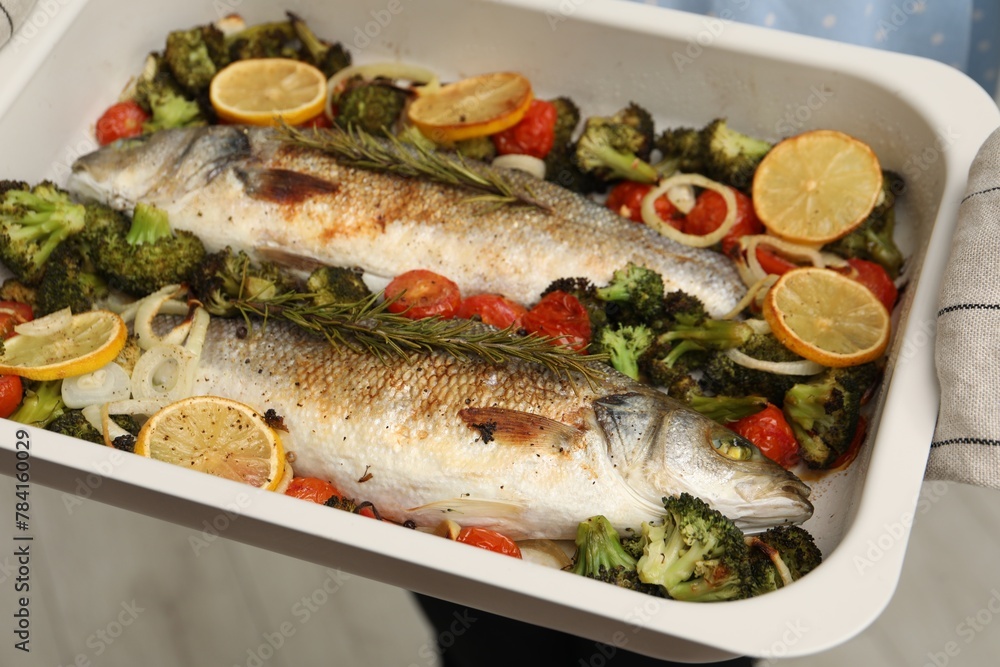 Woman holding baking dish with delicious fish and vegetables indoors, selective focus Woman holding baking dish with delicious fish and vegetables indoors, selective focus