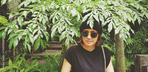 Asian woman is smiling, wearing sunglass and wool hat sitting on wooden bench under tree with green plant background. Girl in casual dress style in vintage tone color with copy space..
