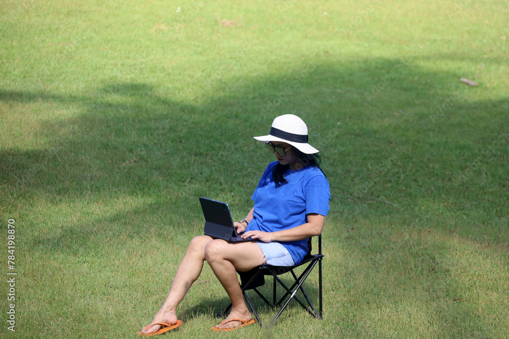 Asian woman in straw beach hat doing online work with computer on folding chair with green grass background, concept of gen z and remote work. Asian woman in straw beach hat doing online work with computer on folding chair with green grass background, concept of gen z and remote work.