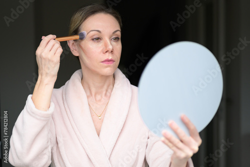 Blonde woman applying makeup with a brush and looking into a mirror