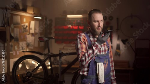 Handsome man holding smartphone using voice command recorder, assistant or takes calling standing in repair bicycle shop