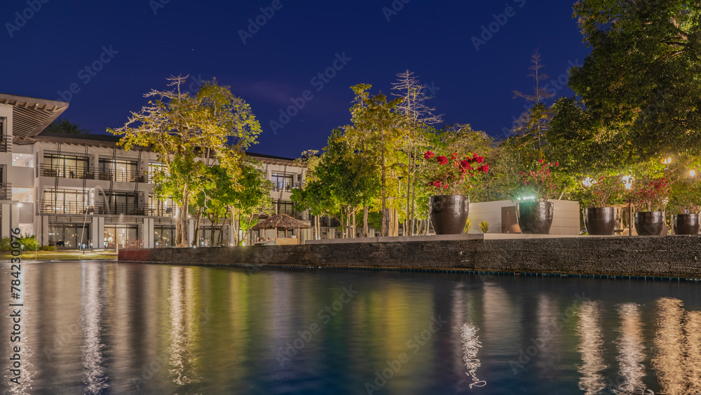 A night in the tropics. Potted flowers bloom by the pool, green trees grow. Reflection, glare on calm water. Stars in the dark blue sky. Electric lighting with street lights. A building with balconies