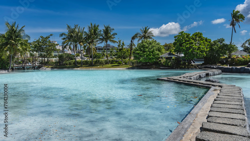 A winding concrete footpath crosses the turquoise water swimming pool. Cottages can be seen in the distance among the green tropical vegetation. Palm trees against a blue sky and clouds. Philippines. 