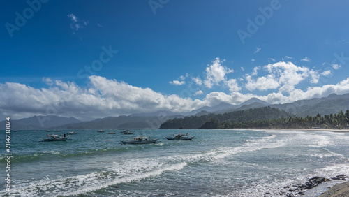 Several traditional Filipino double-outrigger dugout bangka boats are anchored in the ocean. Turquoise waves foam and spread over the beach. Hills against a  blue sky and clouds. Philippines. photo