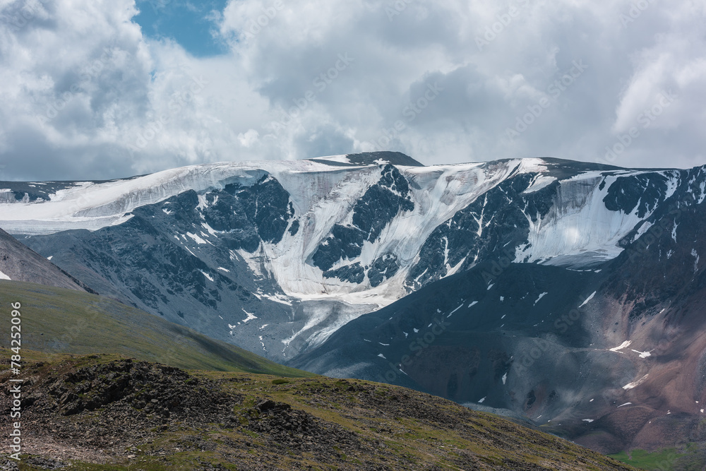 Dramatic view to large snow-capped mountain range in sunlight under gray cloudy sky. Atmospheric landscape with glacier on snowy rocky mountain wall. Shadows of clouds on rocks and high snow mountains